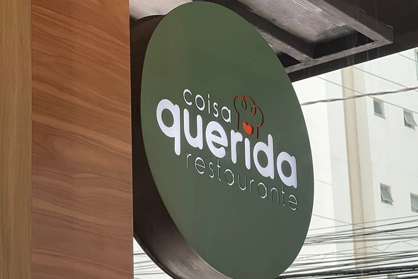 Placa redonda do restaurante Coisa Querida com fundo verde, exibindo o nome do estabelecimento em letras brancas.