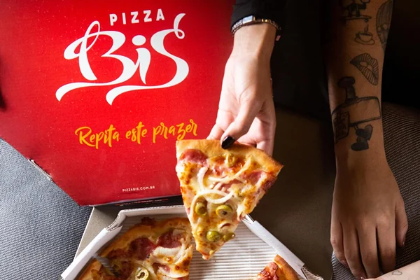 Caixa de pizza da Pizza Bis, com uma fatia sendo servida. A caixa exibe o slogan “Repita este prazer”.