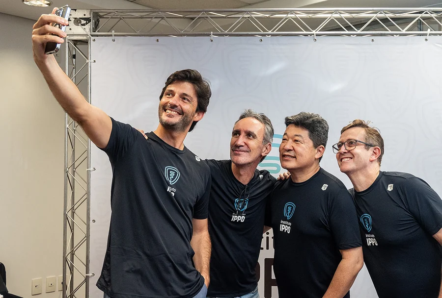 Quatro homens sorridentes do IPPO, vestindo camisetas pretas com logo, fazendo selfie com estrutura metálica e fundo branco.