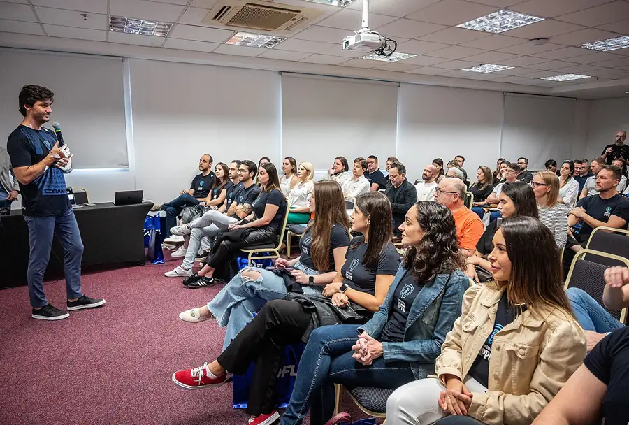 Homem do IPPO falando em microfone para público sentado, sala com carpete vermelho, cadeiras, projetor no teto e janelas