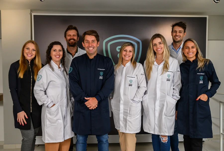 Equipe de profissionais do Instituto IPPO vestindo jalecos brancos e azuis, reunidos em frente a um painel com o logotipo do IPPO, todos sorrindo para a câmera.