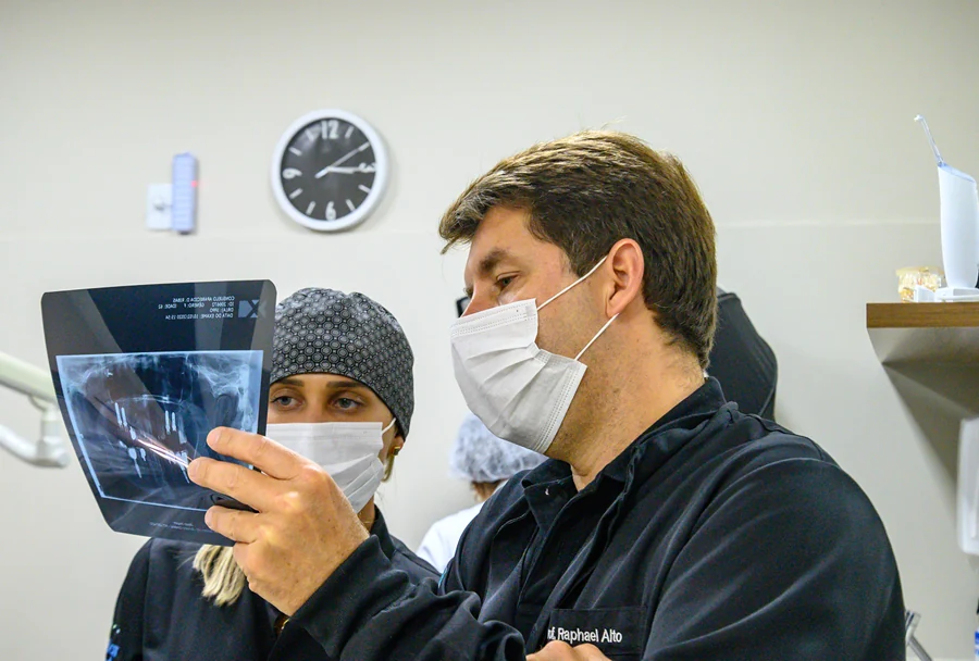 Dentista e assistente examinando um raio-X durante uma consulta odontológica, com relógio ao fundo.