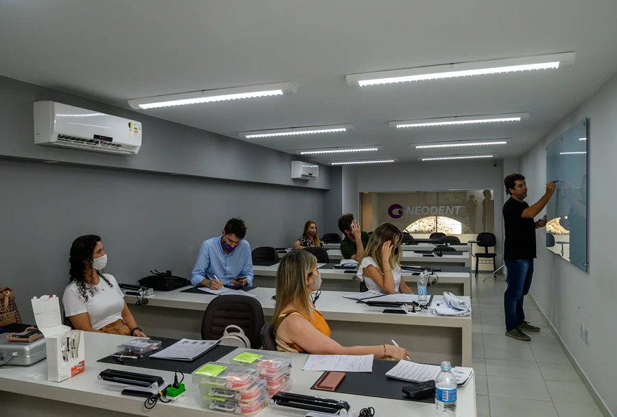 Turma de alunos assistindo aula com professor no quadro branco, em sala de aula do Instituto IPPO com o logotipo da Neodent visível ao fundo.