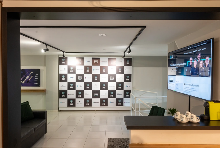 Foto de uma sala de recepção moderna do Instituto IPPO, mostrando uma parede de patrocinadores ao fundo e um monitor exibindo informações institucionais.