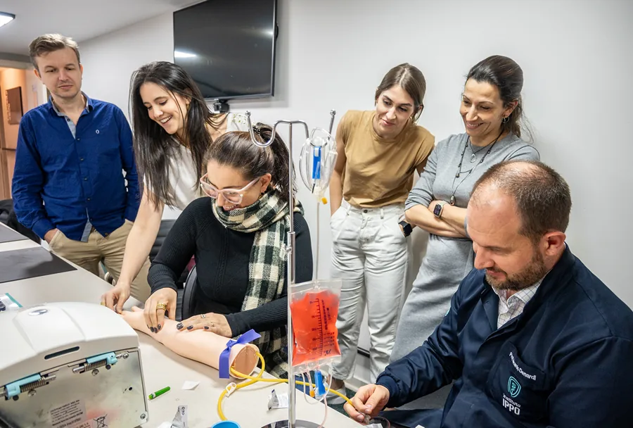 Grupo de alunos observando e interagindo com uma demonstração prática de coleta de sangue, supervisionada pelo professor Ricardo Demétrio.