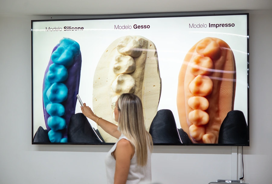 Imagem comparativa de três modelos de dentes (silicone, gesso e impresso) exibida na tela, com a instrutora apontando para o modelo de gesso.