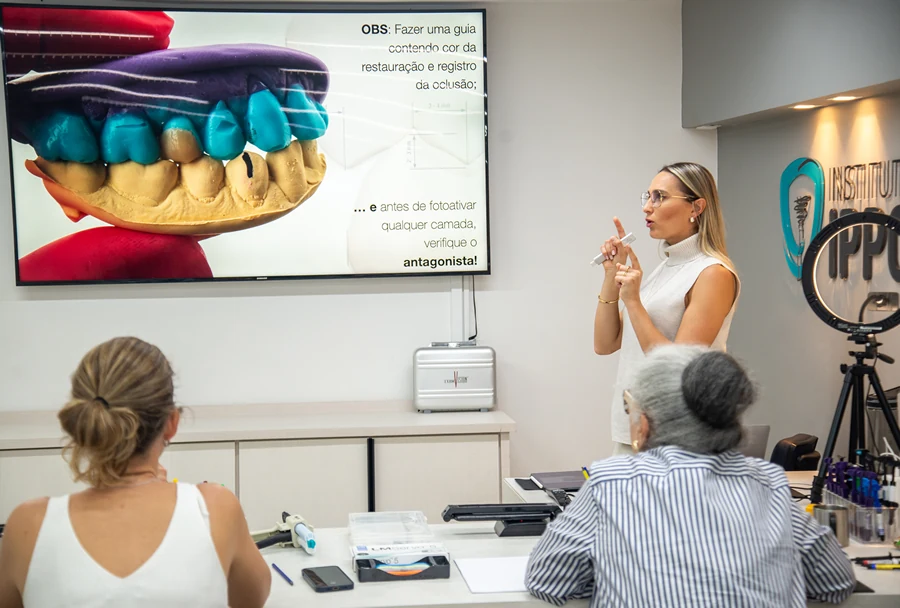 Instrutora apontando para um modelo colorido de arcada dentária projetado em uma tela, explicando sobre restauração e registro da oclusão.